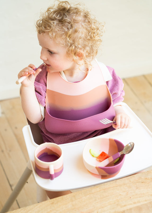 Overhead view of curly-haired toddler girl self-feeding in highchair with Grech & Co Blossom Ombre pink suction bowl, cup and silicone bib on wooden floor — baby led weaning at Milimilu
