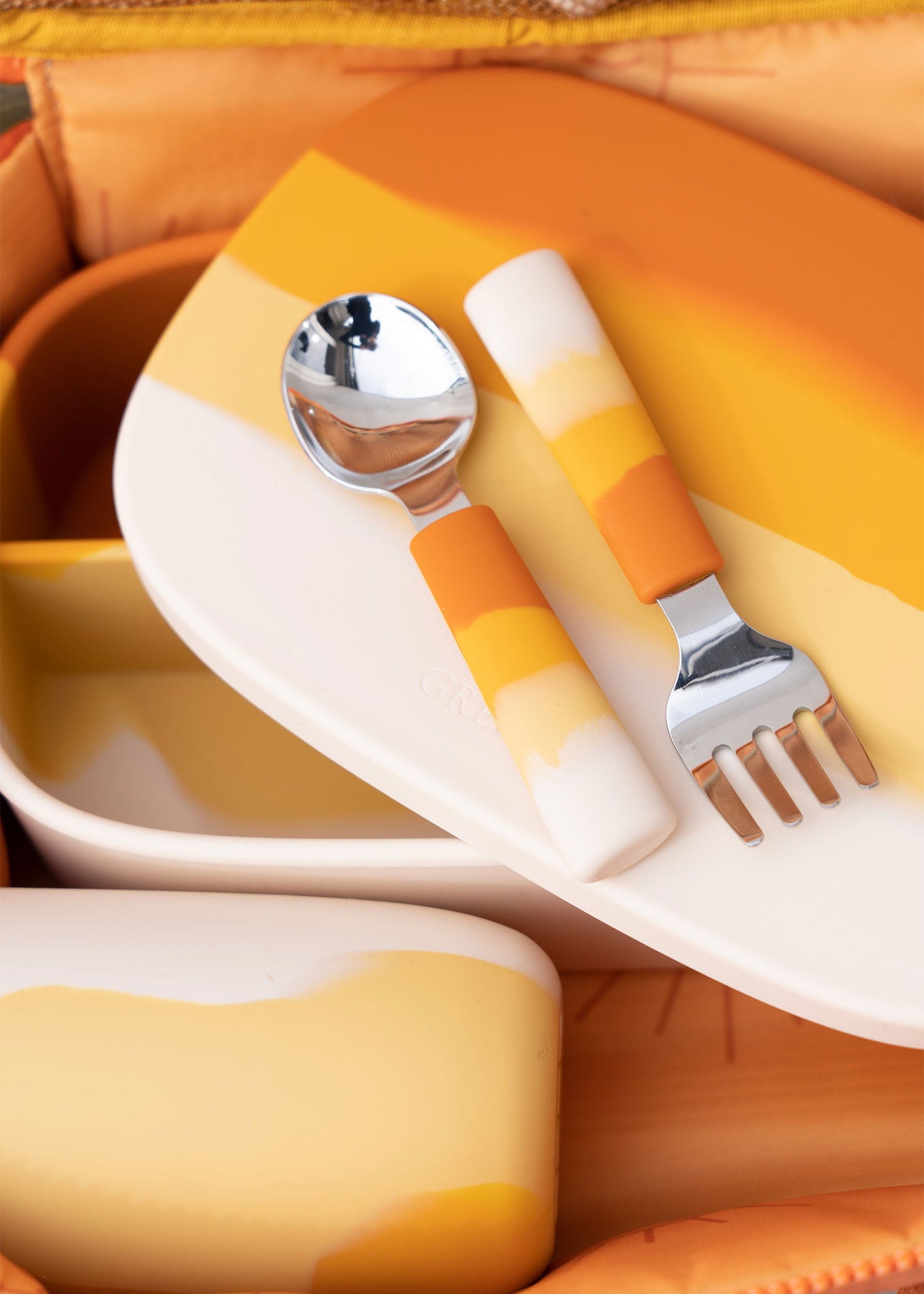 Close-up of Grech & Co Sienna Ombre orange cutlery set spoon and fork resting on matching suction plate and lunchbox inside orange bag — baby led weaning tableware at Milimilu