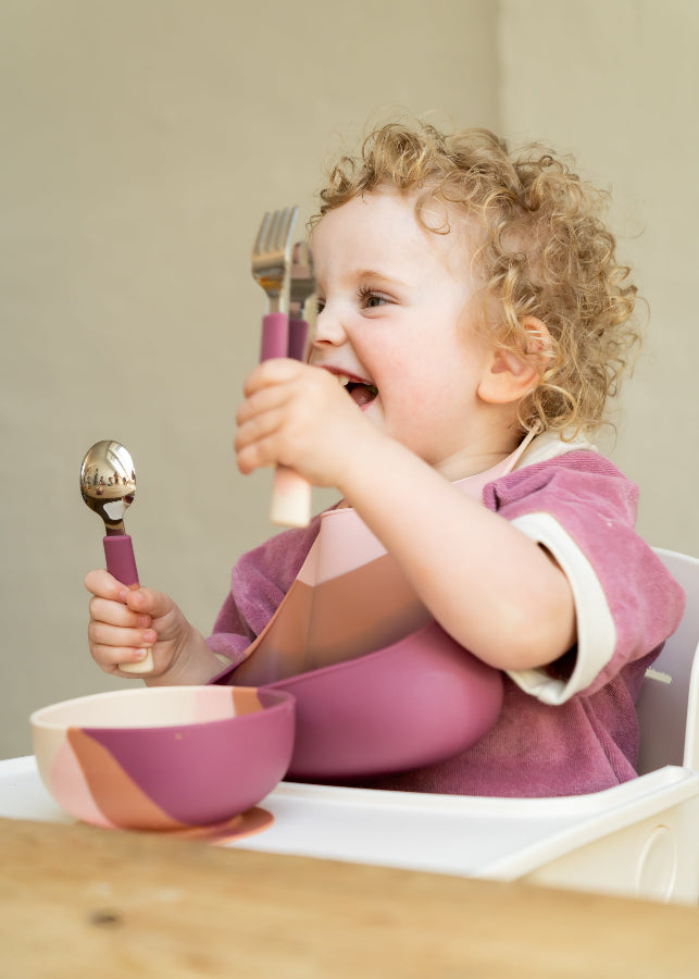 Curly-haired toddler girl laughing in highchair holding Grech & Co Blossom Ombre pink cutlery with matching suction bowl and silicone bib — baby led weaning tableware at Milimilu
