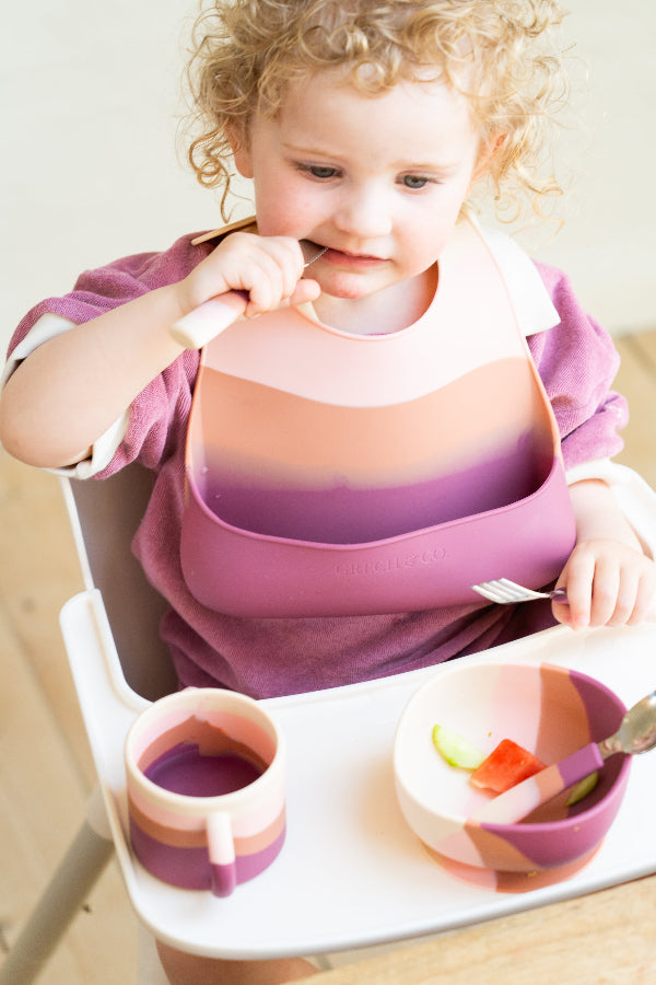 Curly-haired toddler girl in highchair eating from Grech & Co Blossom Ombre pink suction bowl with matching cup and silicone bib — sustainable kids mealtime set at Milimilu