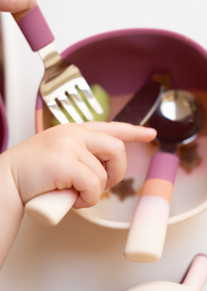 Close-up of baby hand reaching into Grech & Co Blossom Ombre pink suction bowl with fork and spoon inside showing deep mauve interior — eco-friendly toddler mealtime at Milimilu
