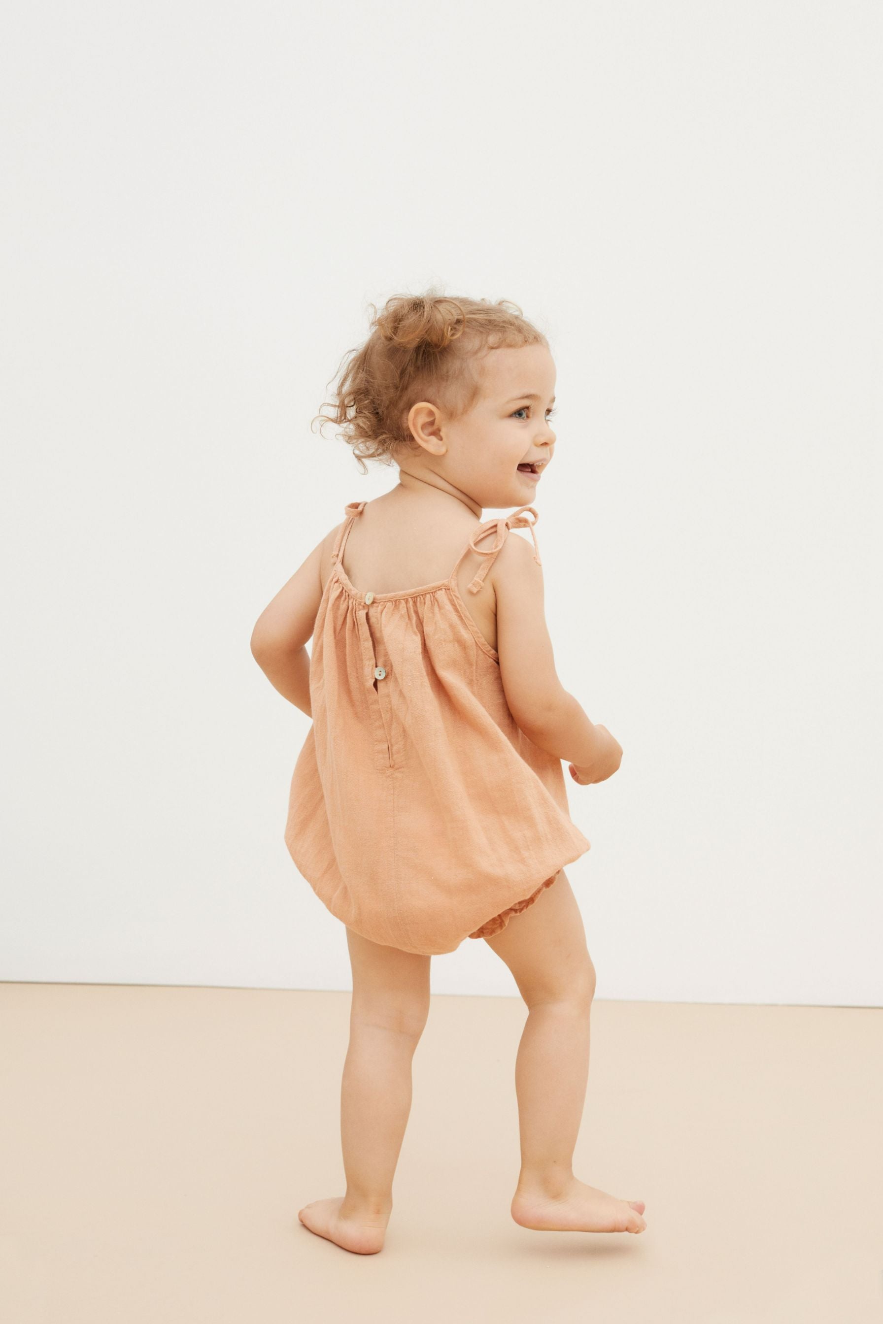 A light peach organic cotton baby girl summer romper is the most adorable outfit this summer. The organic cotton baby romper is breathable and made with lightweight extra soft organic cotton. Have adjustable straps and elasticated legs to fit your baby girl longer.  It is made by The New Society in Portugal. 